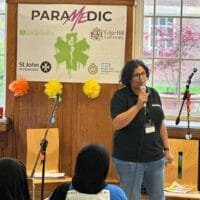 Pooja standing in front of a banner that reads “PARAMEDIC” speaks into a microphone during an indoor event. Colourful paper decorations and microphones on stands are arranged around them, with attendees seated in the foreground and large windows behind the speaker.