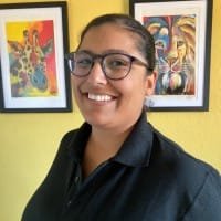 Pooja smiling with glasses stands in front of a yellow wall, wearing a black shirt. Colorful animal art frames are in the background.