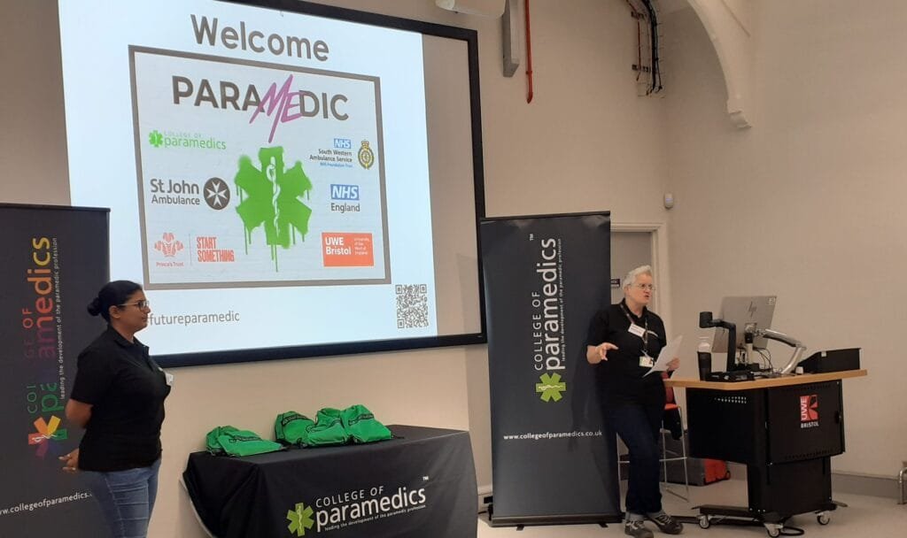 Pooja stands on the left at the front of a lecture room during a PARAMEDIC event. Another speaker stands at a lectern on the right. A large projected slide behind them reads “Welcome PARAMEDIC” and shows logos including the College of Paramedics, NHS England, St John Ambulance, and UWE Bristol. College of Paramedics banners and a table with green bags are positioned at the front of the room.