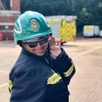 Pooja wearing a green ambulance helmet, dark protective glasses, and a navy protective uniform with reflective yellow stripes poses playfully with one hand raised near her face. Emergency vehicles are blurred in the background of an outdoor training or operational area.