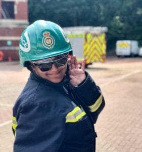 Pooja wearing a green ambulance helmet, dark protective glasses, and a navy protective uniform with reflective yellow stripes poses playfully with one hand raised near her face. Emergency vehicles are blurred in the background of an outdoor training or operational area.