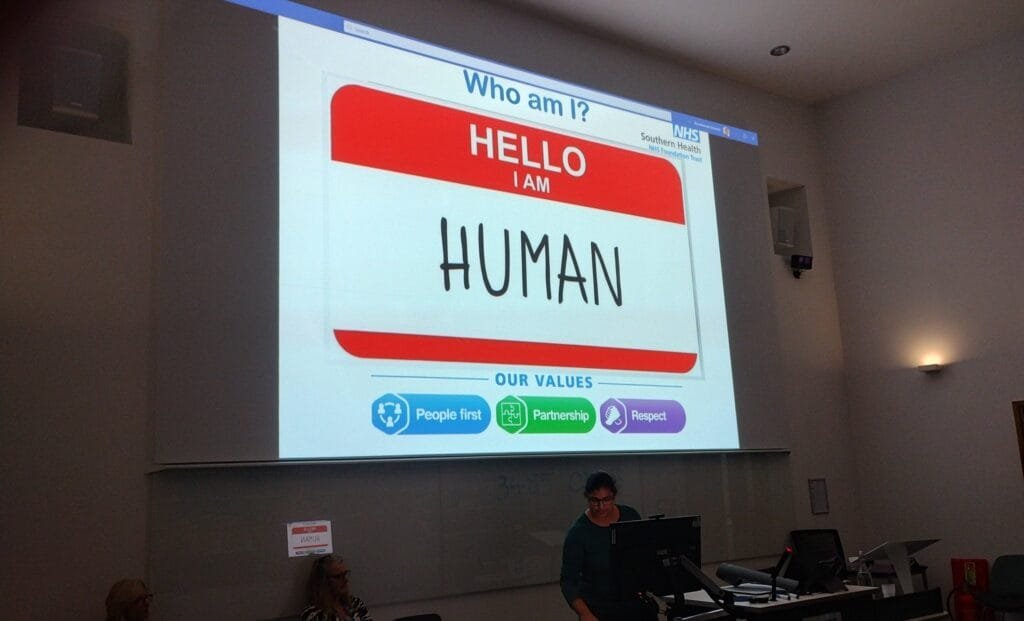 Pooja stands at a lectern presenting in a university lecture theatre. A large projected slide behind her reads “Who am I? Hello I am Human” and displays NHS values including People first, Partnership, and Respect. Several people are seated behind her on the stage, and Bournemouth University branding is visible at the front of the room.