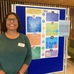 Pooja smiling wearing glasses and a green top stands beside a large academic poster titled “Facilitating inclusive student placements: How can we be part of positive change?” The colourful poster includes sections on best practice, key questions, root causes, future design, and next steps. Pooja is wearing a name badge and standing in a modern indoor space with wooden panelling and seating in the background.