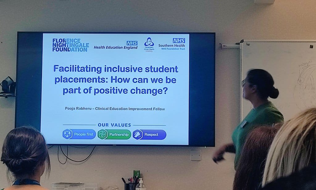 Pooja stands at the front of a room presenting to an audience beside a large screen. The slide reads “Facilitating inclusive student placements: How can we be part of positive change?” and lists Pooja Rabheru as Clinical Education Improvement Fellow, with NHS and Florence Nightingale Foundation logos and values icons including People first, Partnership, and Respect.